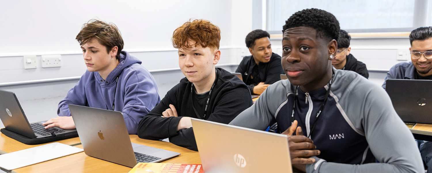 Three male students sit in class side by side, each looking at a laptop.