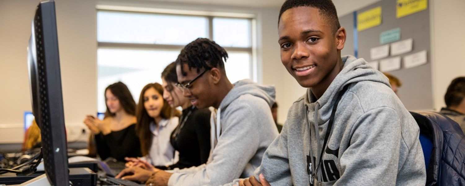 A male student sits at a computer and smiles to camera.