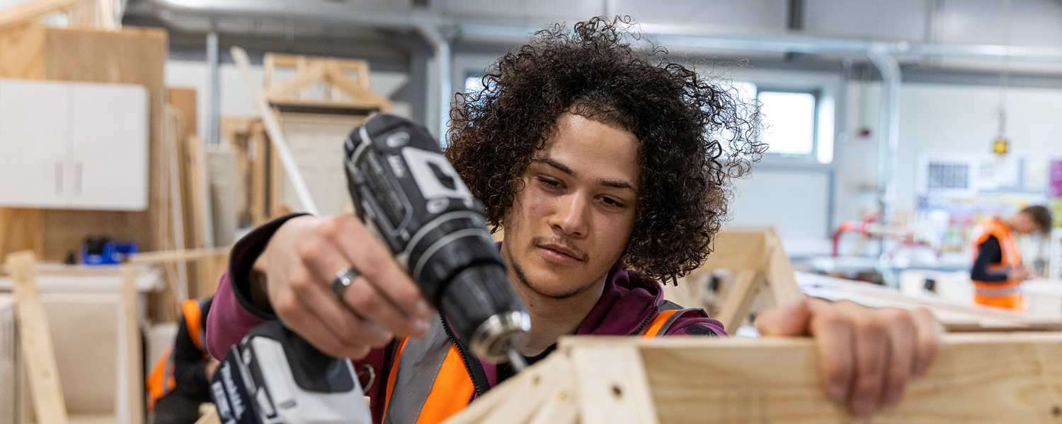 A male Construction student works on a carpentry project using a drill.