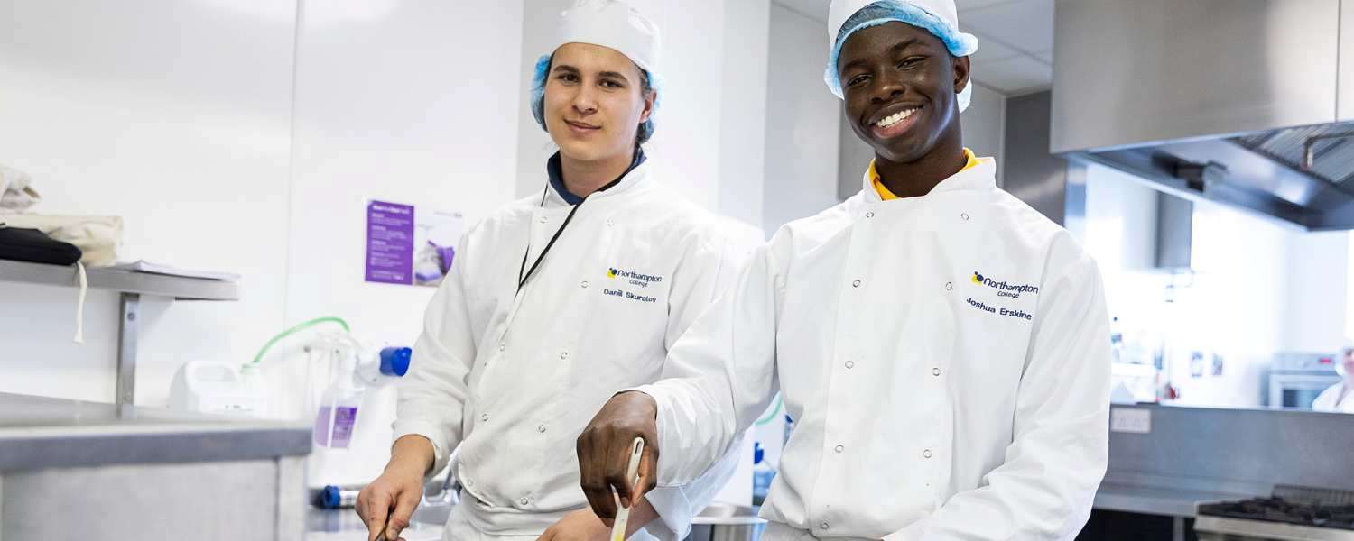 Two male student posse in the College kitchen wearing chef whites.