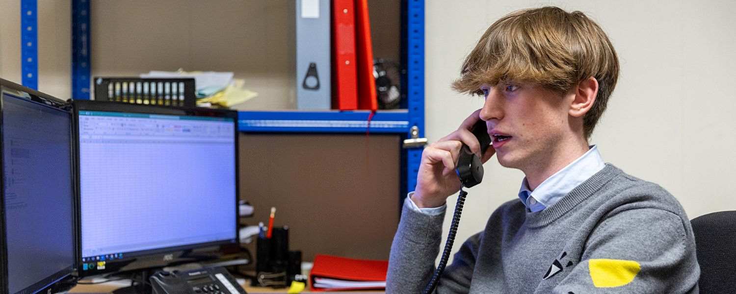 A male apprentice sits at a computer and speaks to a customer on the phone.