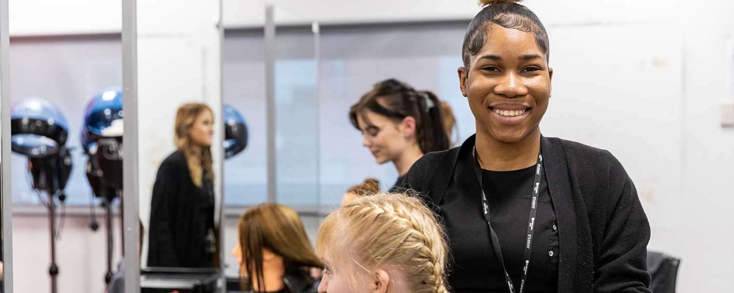 Student smiling to camera, braiding a customers' hair.