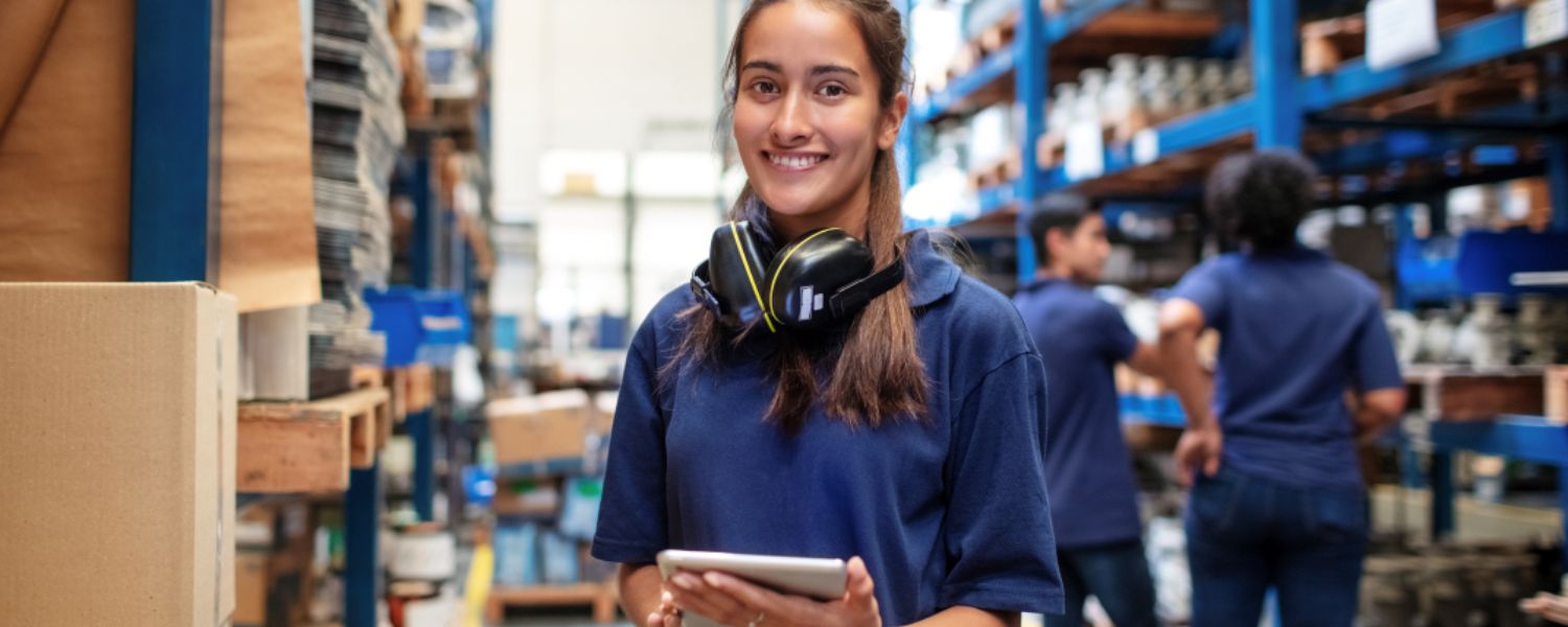A young female poses with a tablet in a logistics warehouse, smiling to camera.