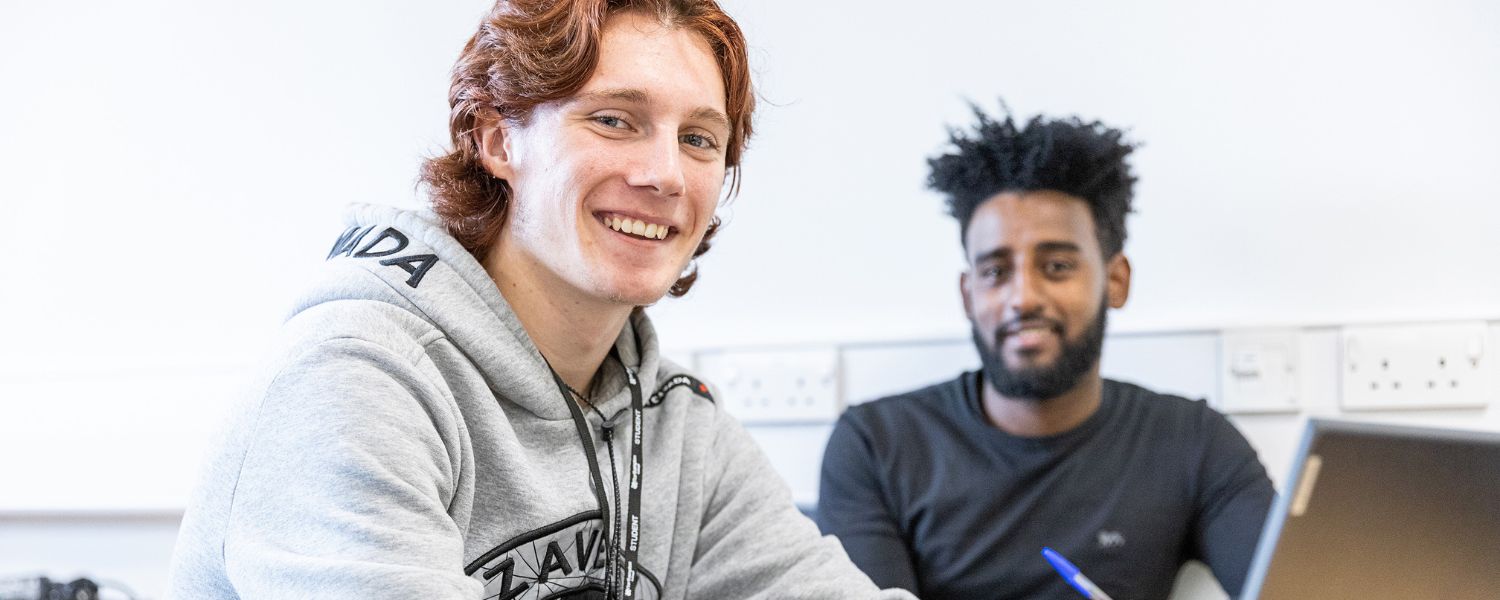 Two male students sit side by side and smile to camera.