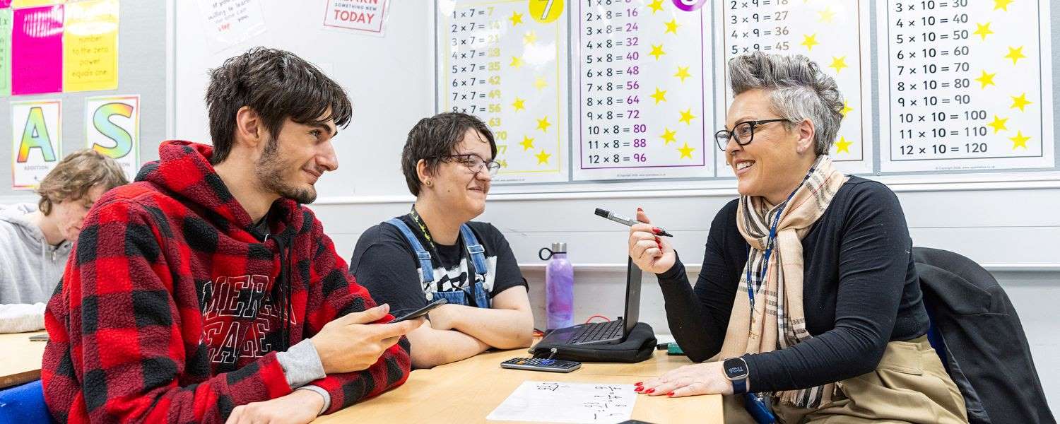 A female teacher sits opposite two students (one male, one female) in a maths classroom. The students are following along on their worksheet while the teacher helps them.