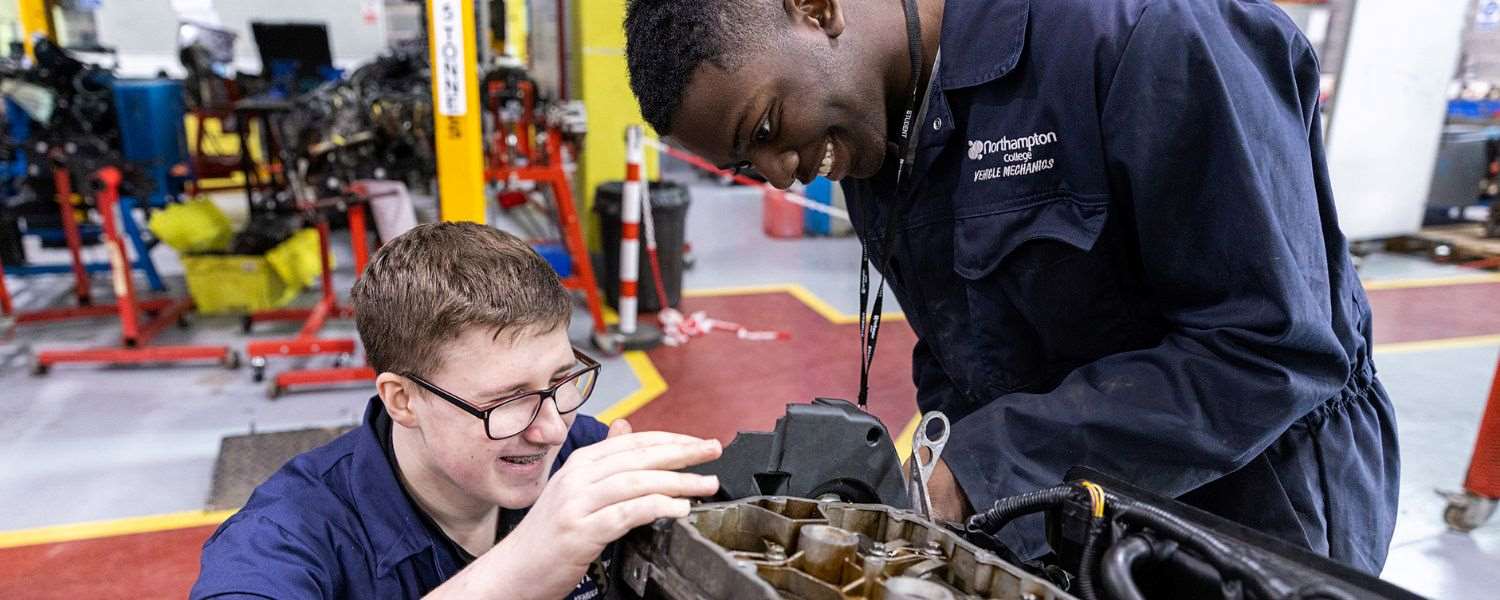Two male students in blue overalls work on a car in our vehicle workshop.