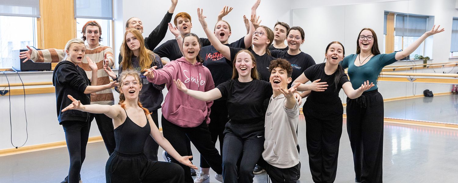 A group of performing arts students pose and smile to camera, holding their arms in the air.