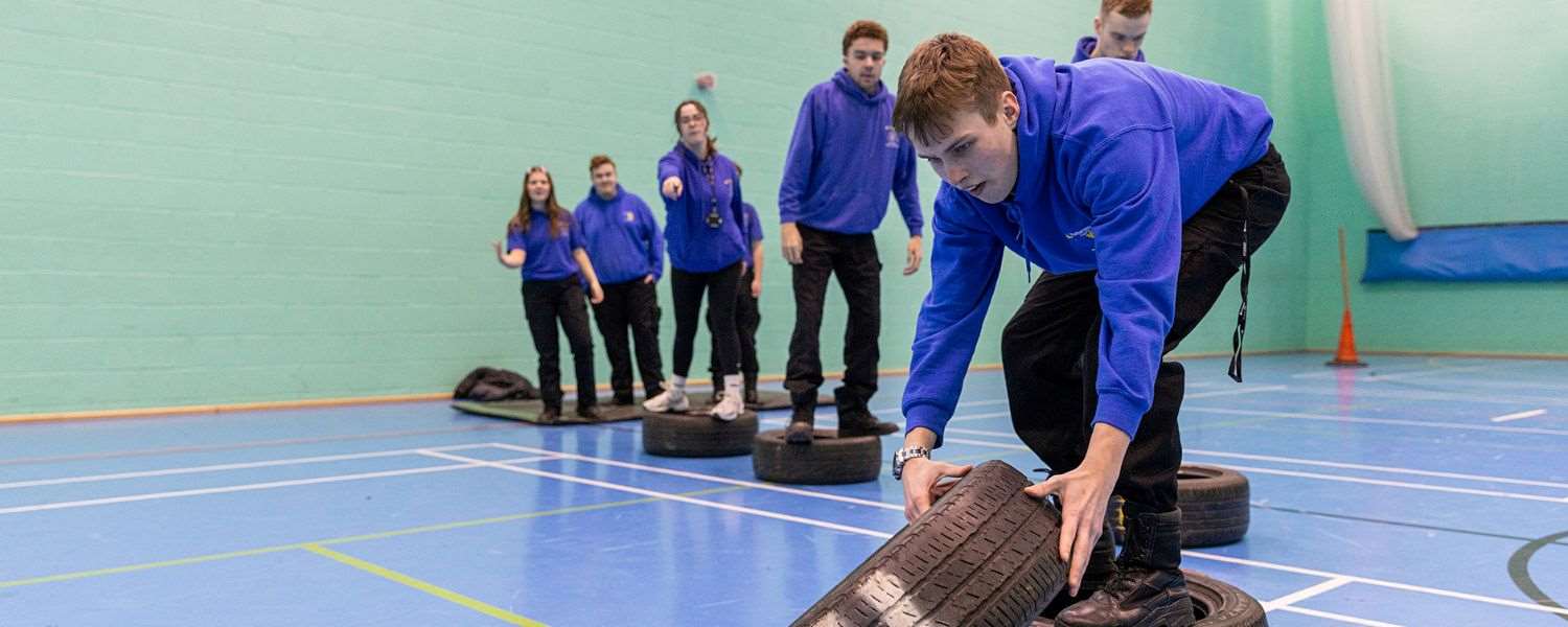 A group of students in NC Public Services uniform take part in a practical exercise in the sports hall. A male student is shown flipping a tyre.
