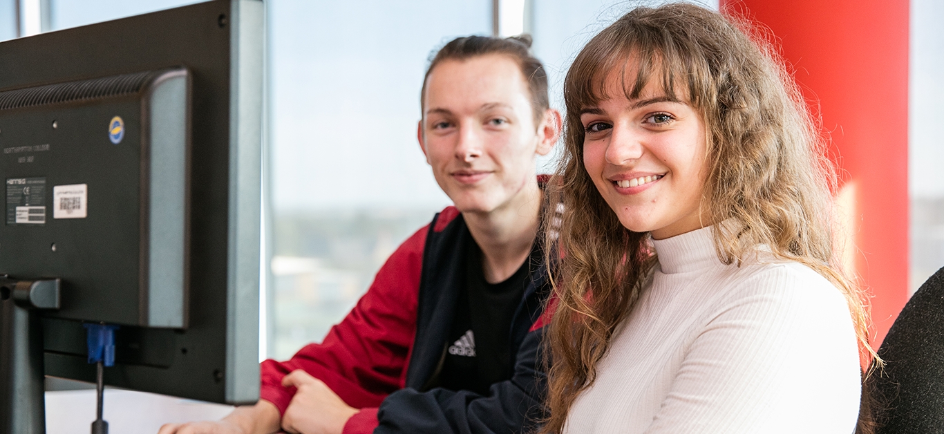 Two students sat at computer smiling.