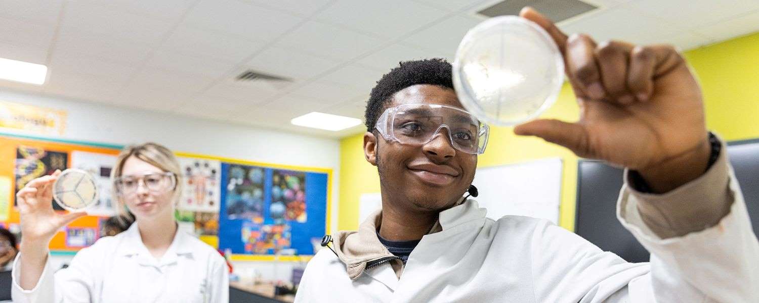 A male and a female student, wearing white lab coats and goggles, both hold a petri dish to camera.