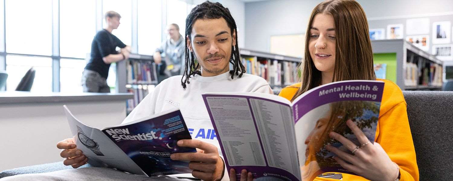 Two students, a male and a female, read side by side in a library. The male is reading The New Scientist and the female is reading a wellbeing textbook.