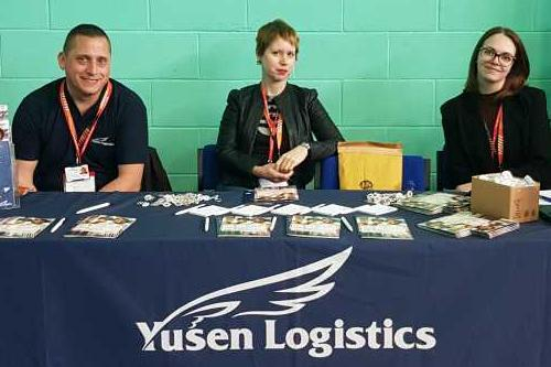 Image shows visiting employers from Yusen Logistics sat behind a desk at a careers fair, smiling to camera.