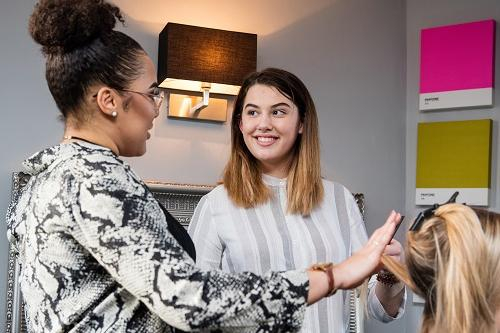 Hairdressing student on work placement poses with hairdresser while they work together on a female client.