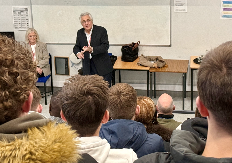 Man standing at the front of the room with students sitting listening. 