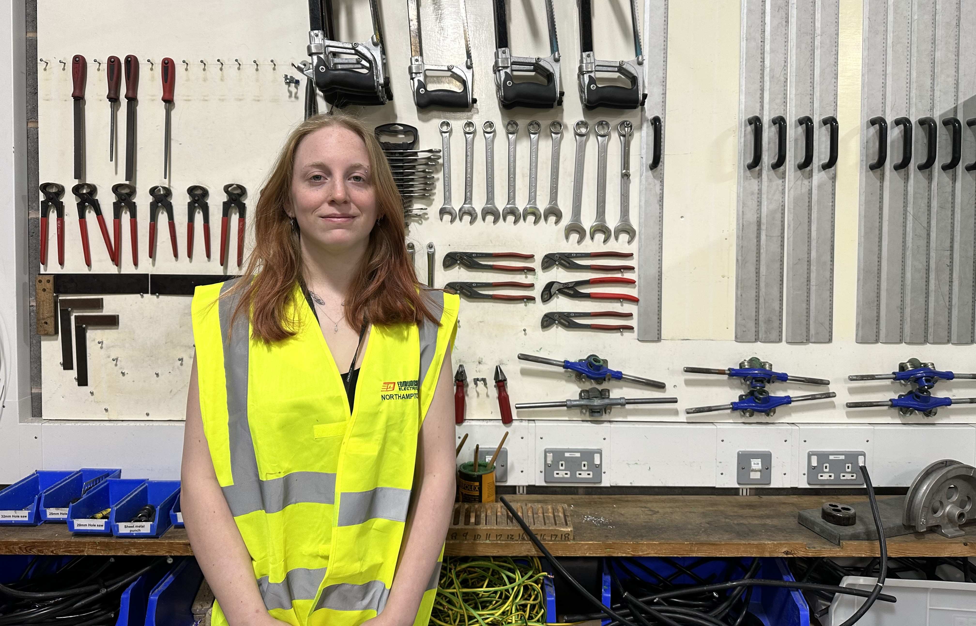 Photo shows apprentice Charlie Kirby, standing against a wall of tools wearing hi-vis and smiling to camera